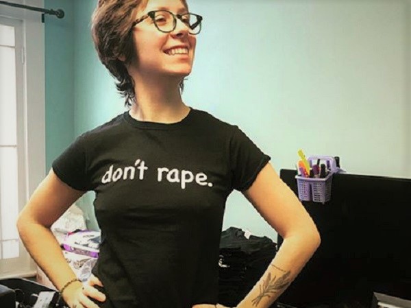Photo of a person from the waist up with their hands on their hips. They have light skin, short brown hair, and dark plastic-framed glasses. They are wearing a black t-shirt with large white printing across the chest reading, "don't rape." light-skinned person from the chest up their arms crossed. They are wearing a black hoodie with white text over the left chest reading, "NEWFRDM." The black hood is up over a grey touque with a white patch on the front. On the white patch is black text reading, "GENDER FREEDOM CLUB."